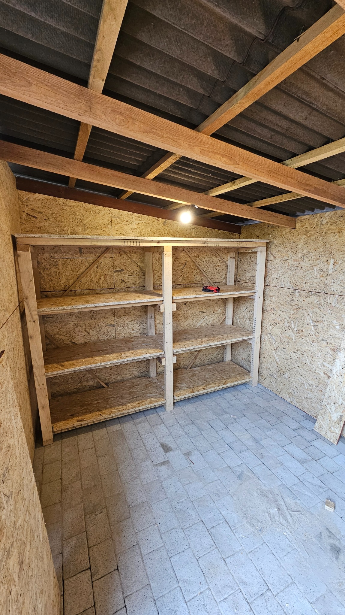 Drewniany regał z półkami z płyty OSB w pomieszczeniu wykończonym płytami OSB i wyłożonym kostką brukową. Na jednej z półek leży narzędzie.