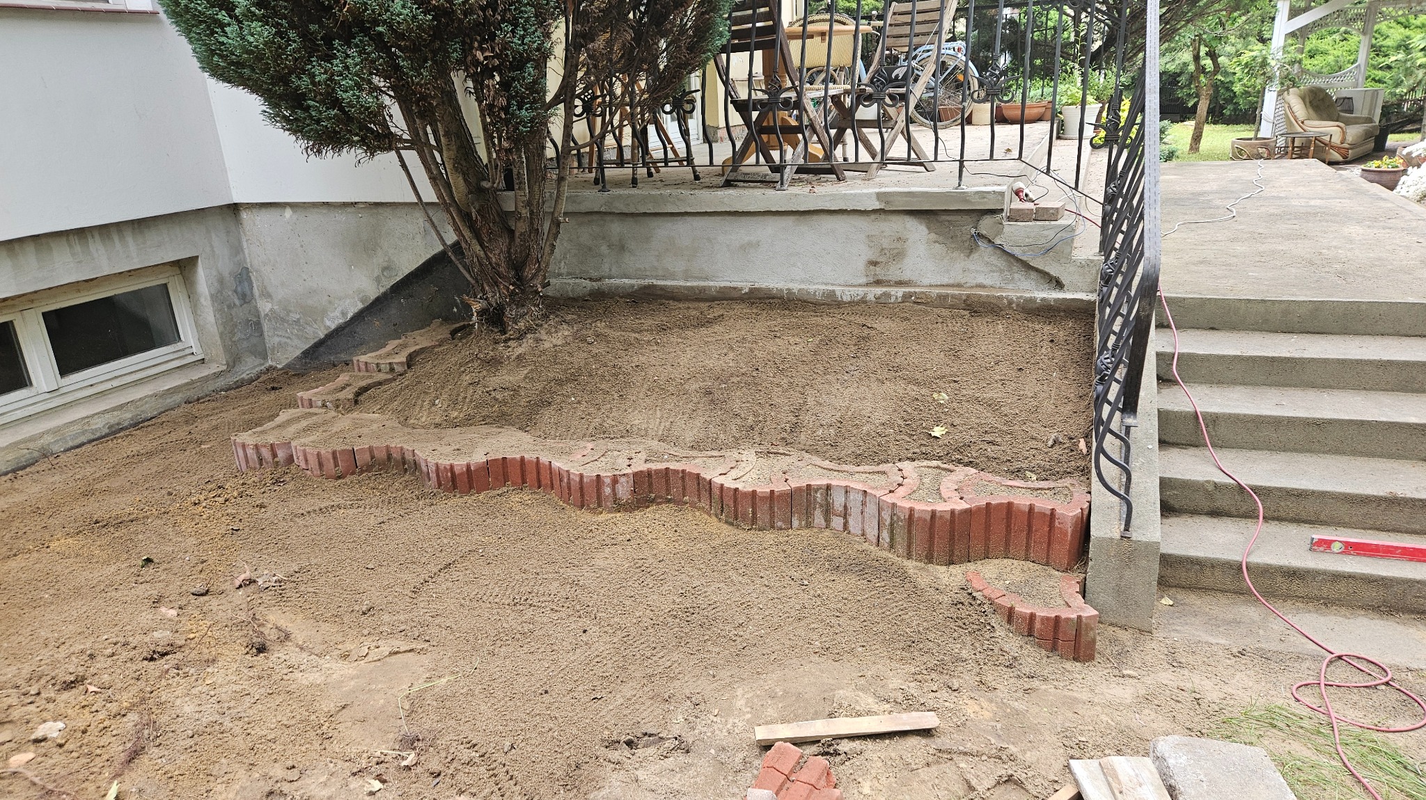 Układanie falistej obwódki z czerwonej kostki brukowej na ziemi przy ścianie budynku i schodach, widoczne narzędzia i kabel.