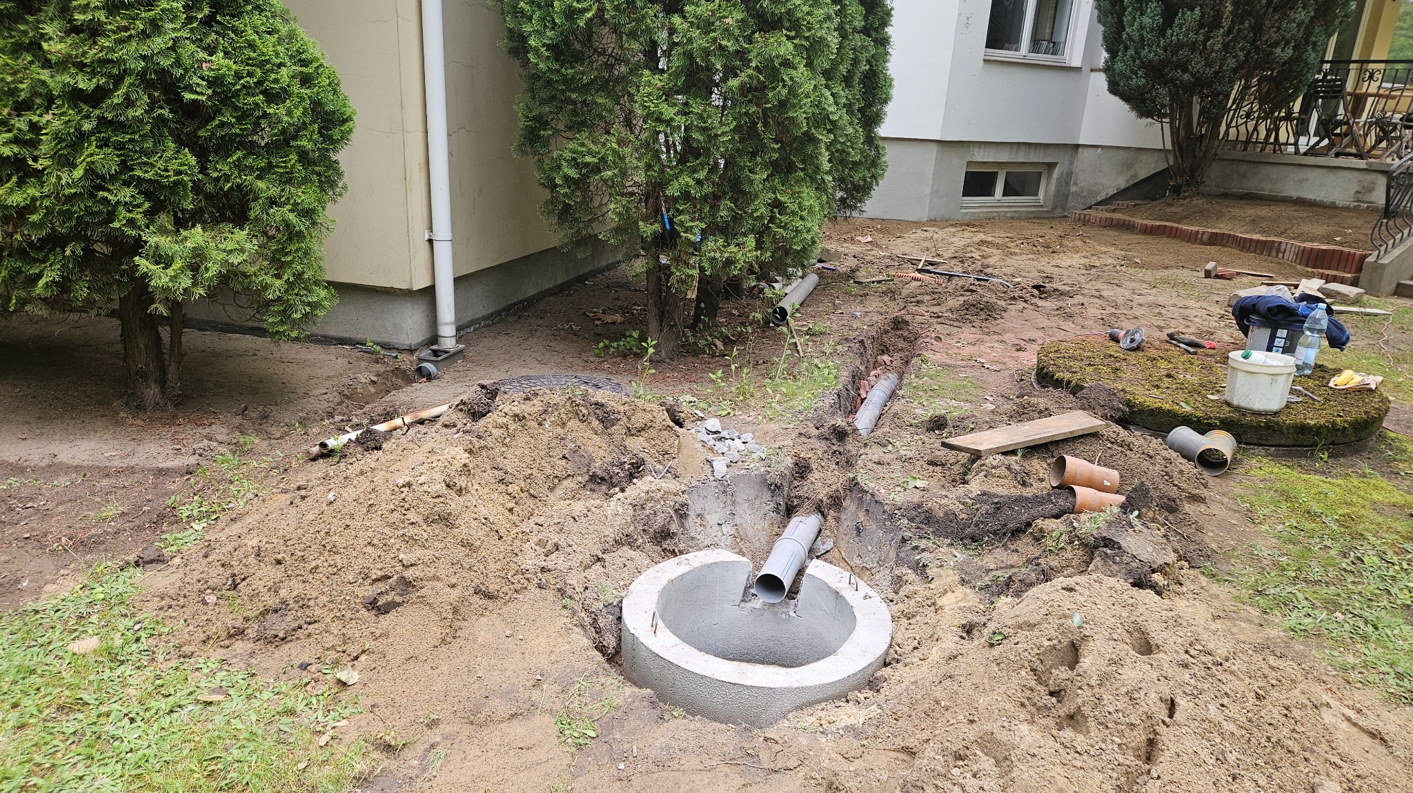 Montaż rur kanalizacyjnych w wykopie na zewnątrz budynku. Widoczny betonowy krąg i rury z tworzywa sztucznego, ziemia oraz narzędzia pracy.