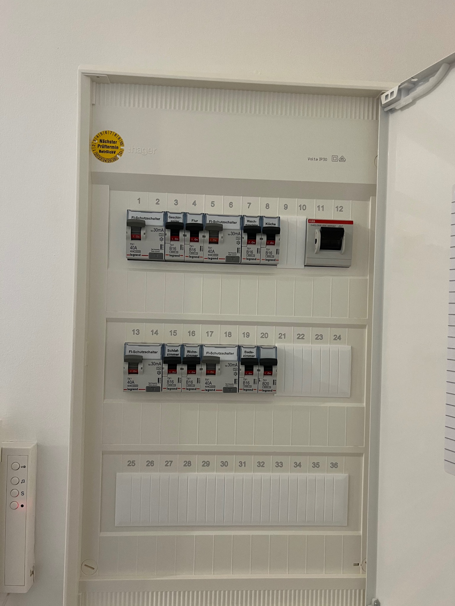 Otwarta skrzynka bezpiecznikowa z widocznymi bezpiecznikami i oznaczeniami, naklejka z datą przeglądu na białej obudowie. Widoczny też fragment domofonu.