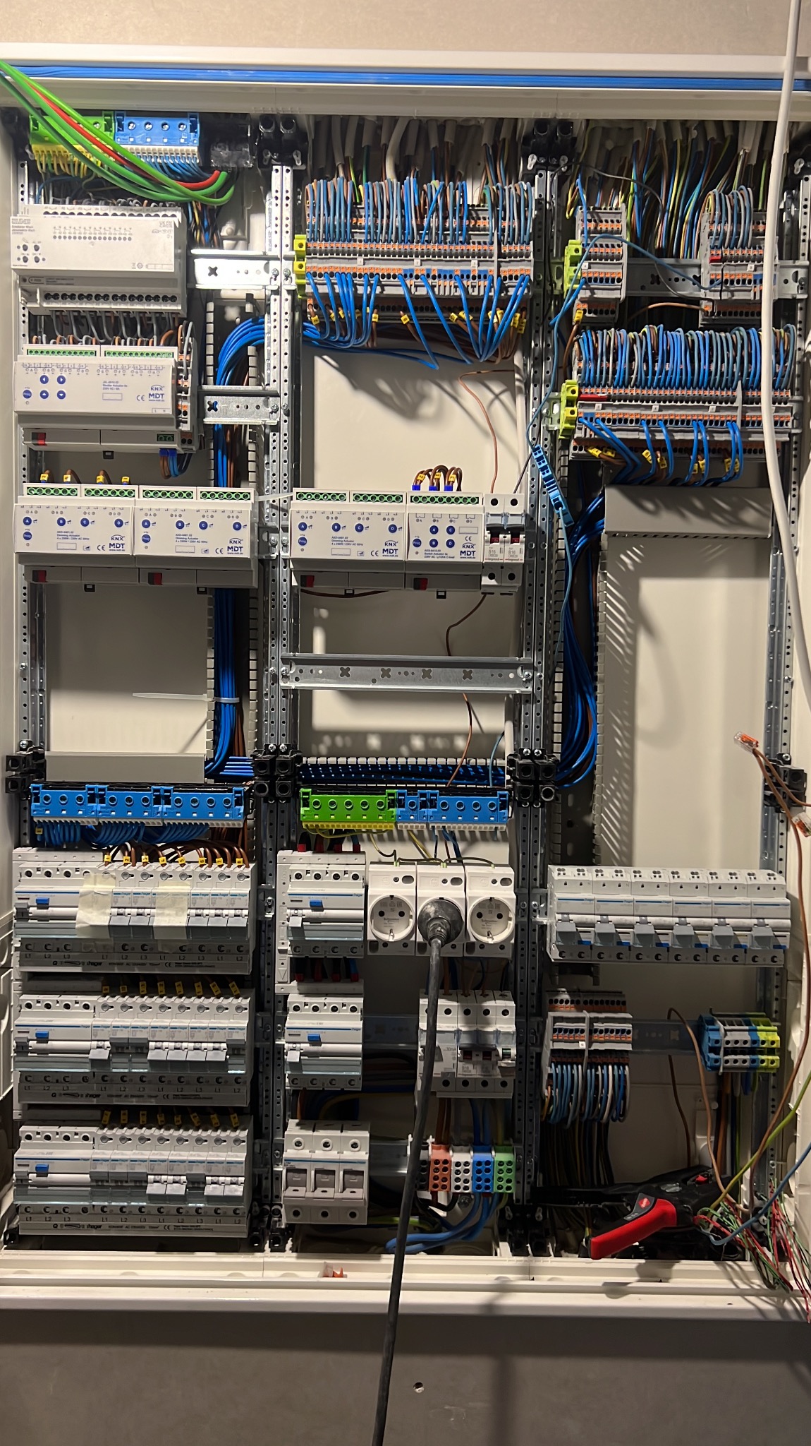 Rozbudowana skrzynka elektryczna z licznymi zabezpieczeniami, przewodami w różnych kolorach i elementami montażowymi. Widoczne gniazdka i narzędzia elektryczne.