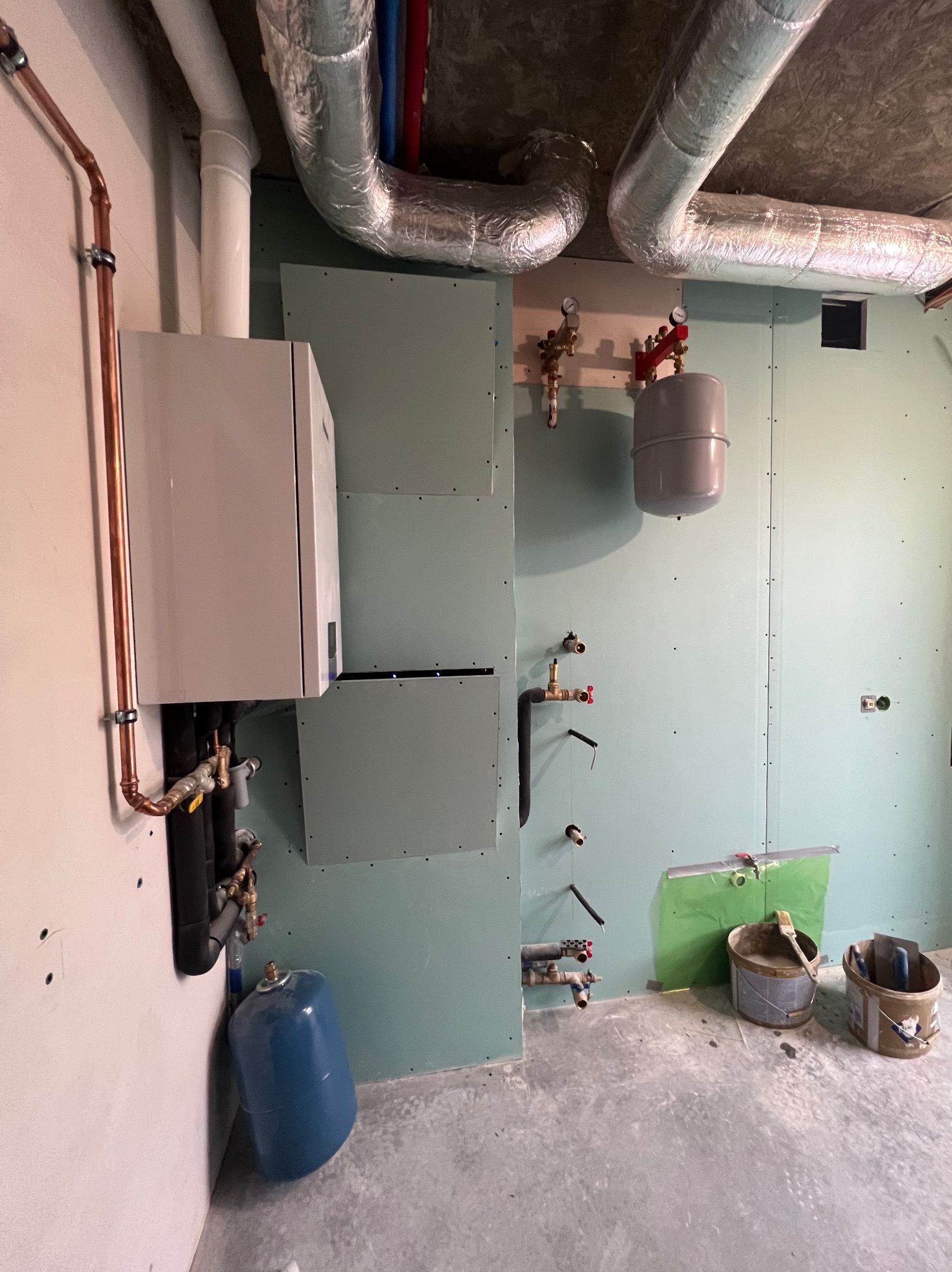 Montaż kotła grzewczego z miedzianymi rurami na ścianie, instalacja wentylacyjna pod sufitem, ściany wykończone płytami gipsowymi, widoczne narzędzia i akcesoria instalacyjne.