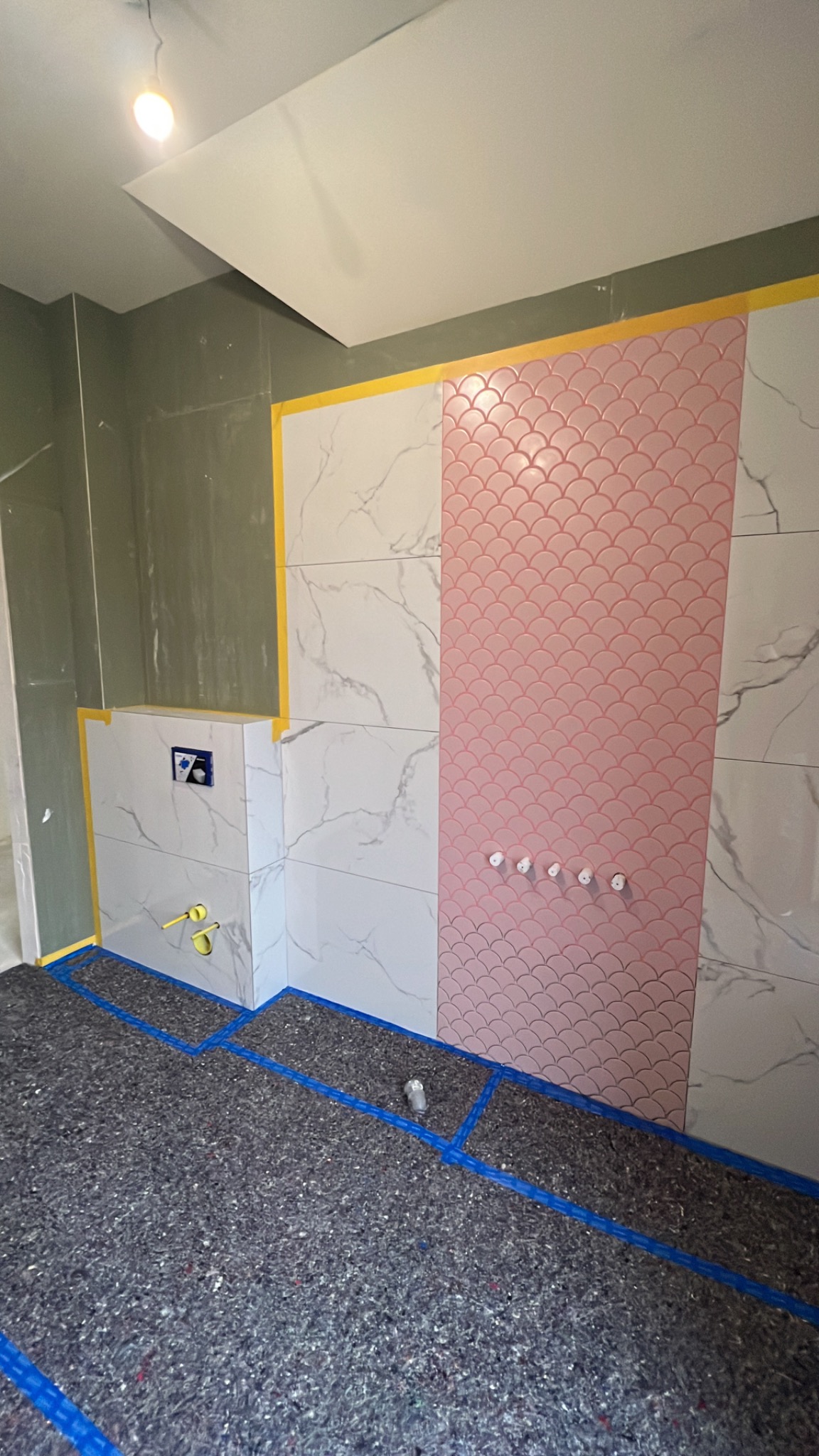 Remont łazienki: biało-różowe płytki w łuski na ścianie, zabezpieczona podłoga, żółta taśma malarska, instalacja podtynkowa WC. Widok na fragment łazienki w trakcie prac.