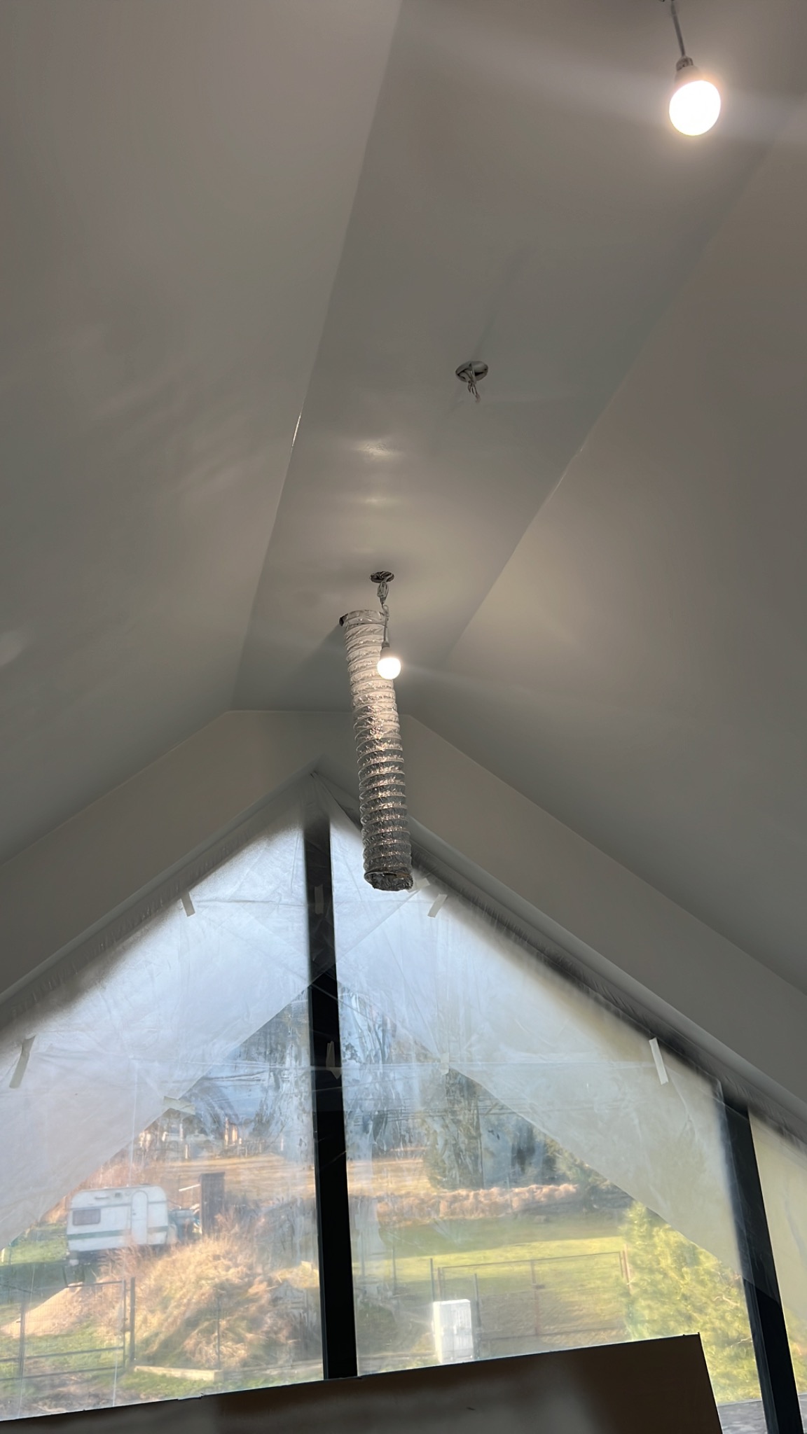 Wnętrze z wysokim stropem i dużym oknem zabezpieczonym folią malarską. Na suficie widoczne lampy, jedna z nich z osłoną w kształcie tuby.