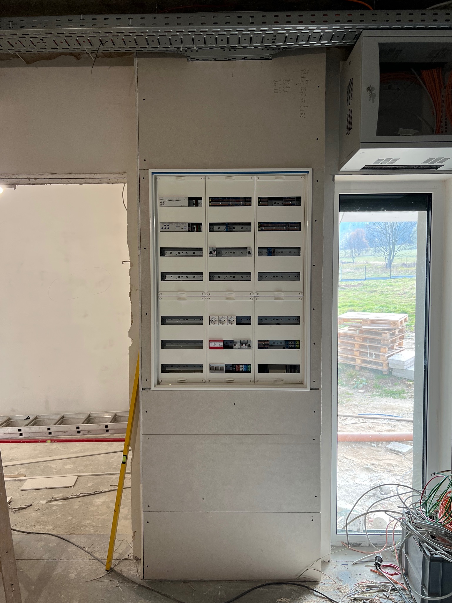 Rozdzielnia elektryczna zamontowana w ścianie z płyt kartonowo-gipsowych, widoczne kable i przewody. W tle drabina i okno z widokiem na zewnątrz. Poziomica oparta o ścianę.