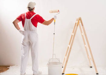 Malarz w białym kombinezonie i czerwonej koszulce maluje ścianę wałkiem. Obok stoi drewniana drabina. Widok z tyłu, jasne oświetlenie.