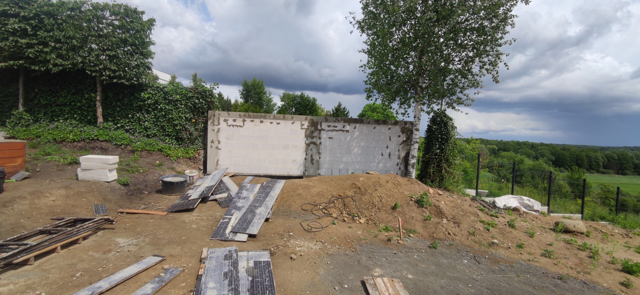 Wznoszenie ścian z białych bloczków na tle zieleni i pochmurnego nieba. Na pierwszym planie materiały budowlane i narzędzia.