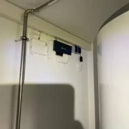 Centrala rekuperacyjna TP-Link w białym pomieszczeniu. Widoczna rura instalacyjna i biały zbiornik. Kompaktowe rozwiązanie z podłączeniami.