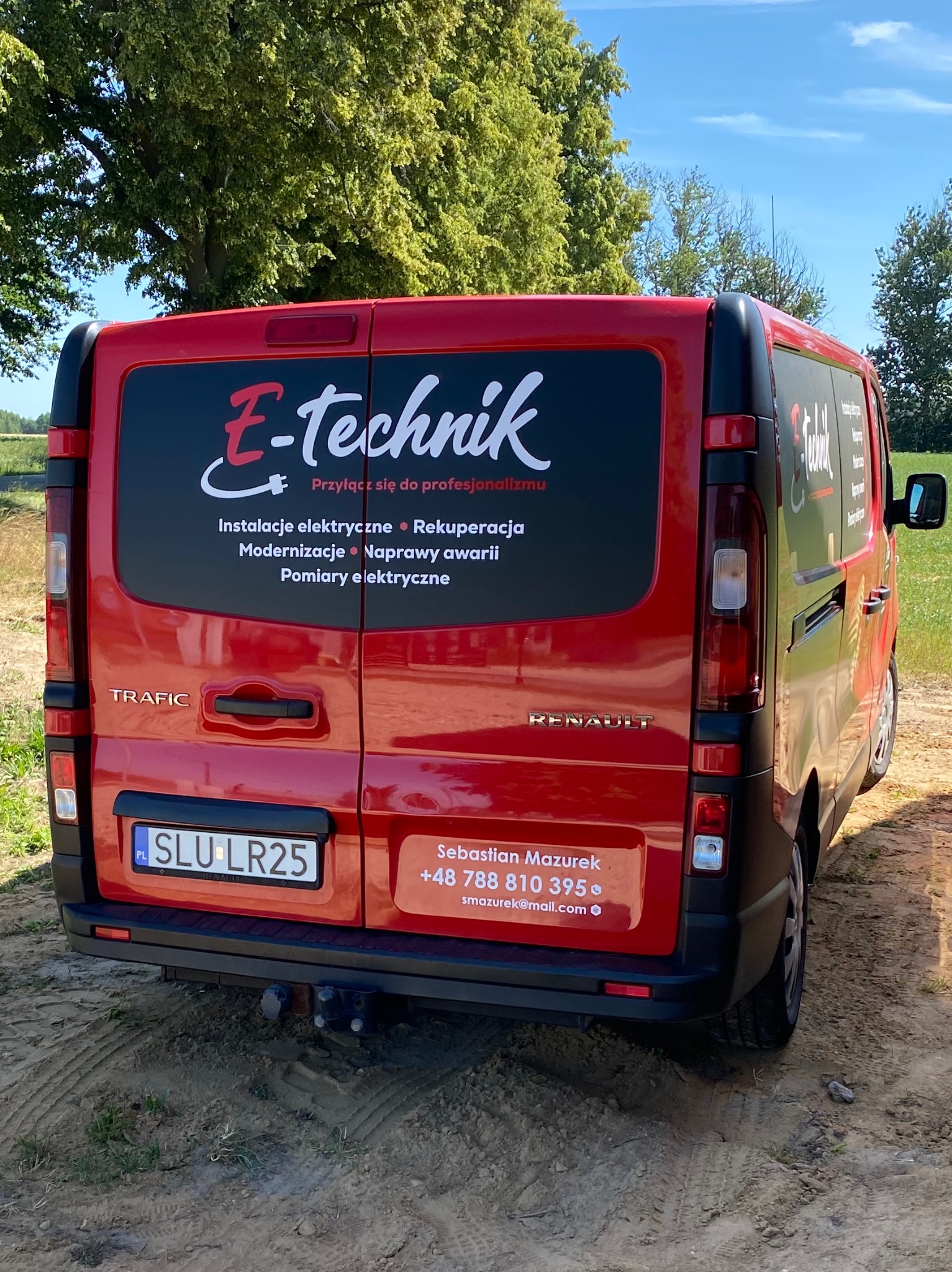Czerwony samochód dostawczy E-technik z reklamą usług elektrycznych i rekuperacji, zaparkowany na polnej drodze. Widoczne dane kontaktowe firmy.