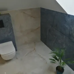 Wnętrze łazienki po remoncie: biała toaleta podwieszana, marmurowe płytki w odcieniach beżu i szarości, roślina doniczkowa. Nowoczesny design z dbałością o detale.