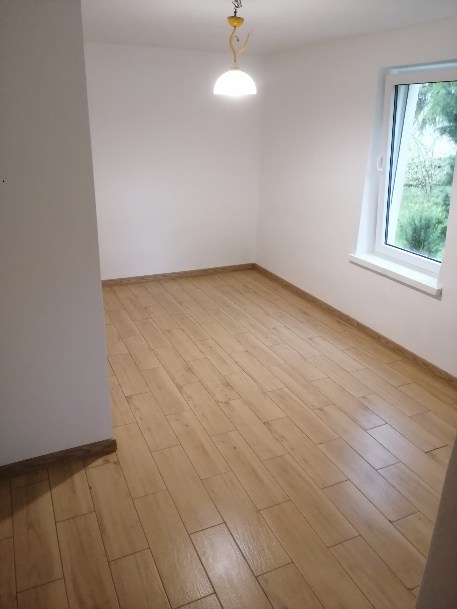 Jasne panele podłogowe ułożone w pustym pokoju z białymi ścianami i oknem. Widoczny fragment lampy sufitowej i listwy przypodłogowe.
