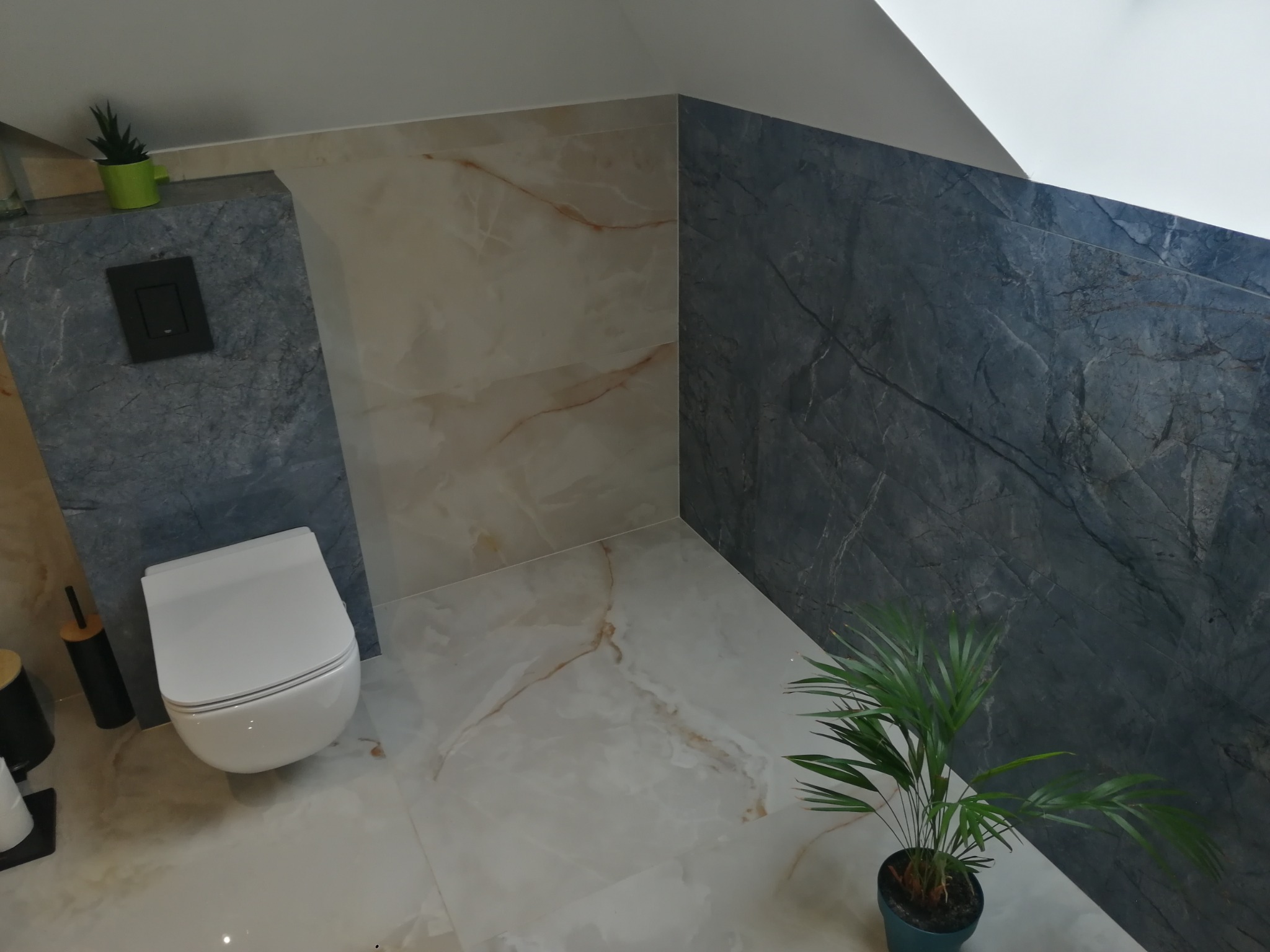 Wnętrze łazienki po remoncie: biała toaleta podwieszana, marmurowe płytki w odcieniach beżu i szarości, roślina doniczkowa. Nowoczesny design z dbałością o detale.