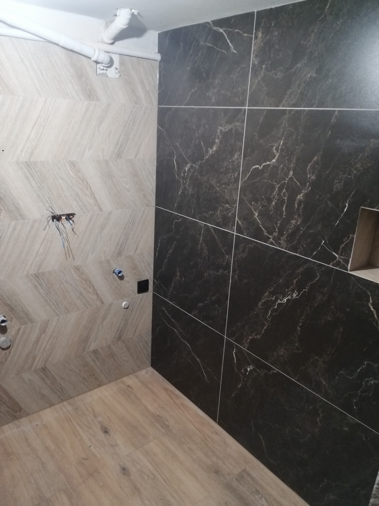 Wnętrze łazienki w trakcie wykańczania: ułożone płytki w dwóch wzorach (chevron i imitacja marmuru), widoczne instalacje hydrauliczne i elektryczne, panele podłogowe.