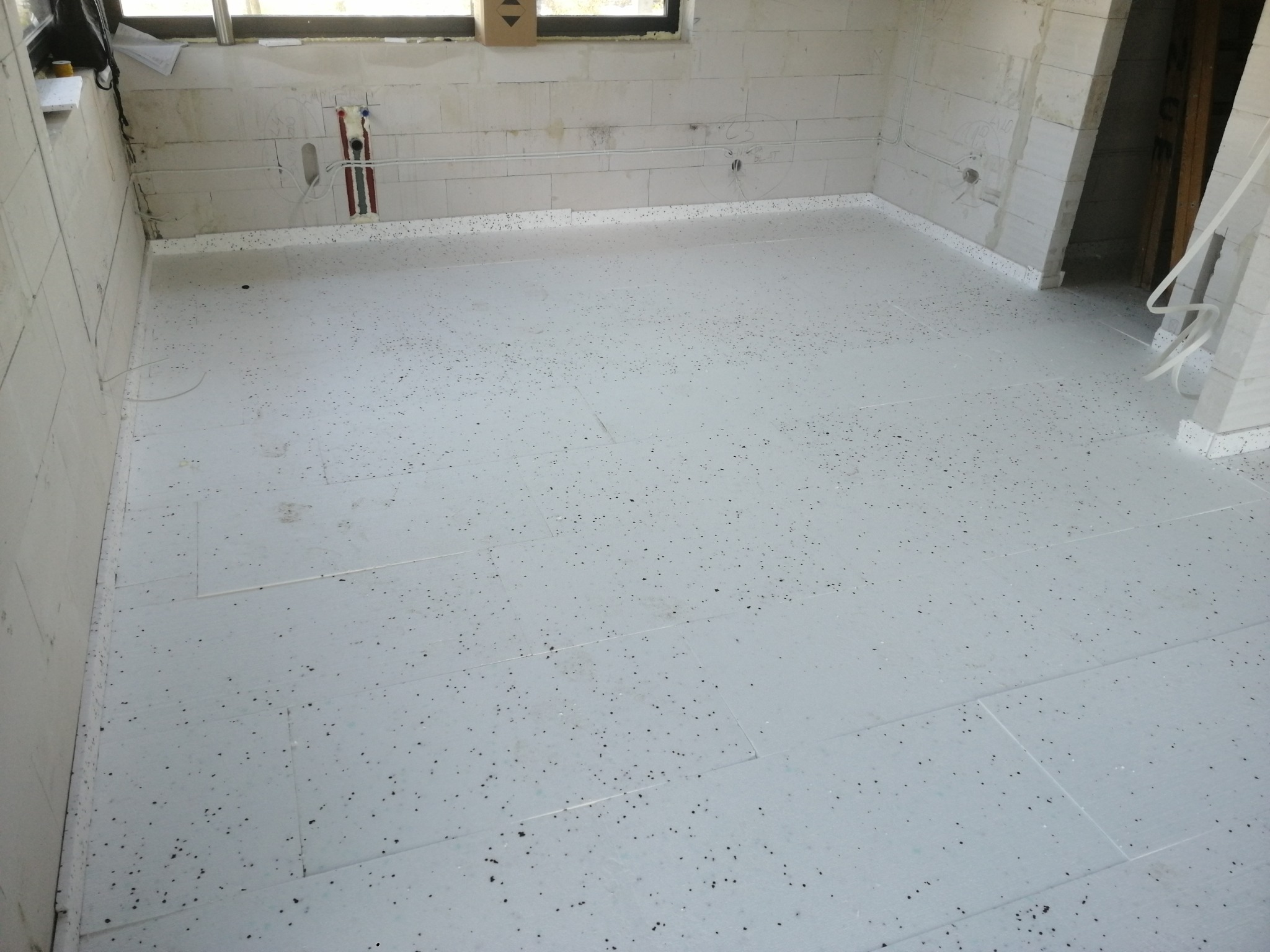 Białe płyty izolacyjne pod wylewkę, ułożone na podłodze w surowym wnętrzu z widocznymi instalacjami. Przy oknie stoi karton.