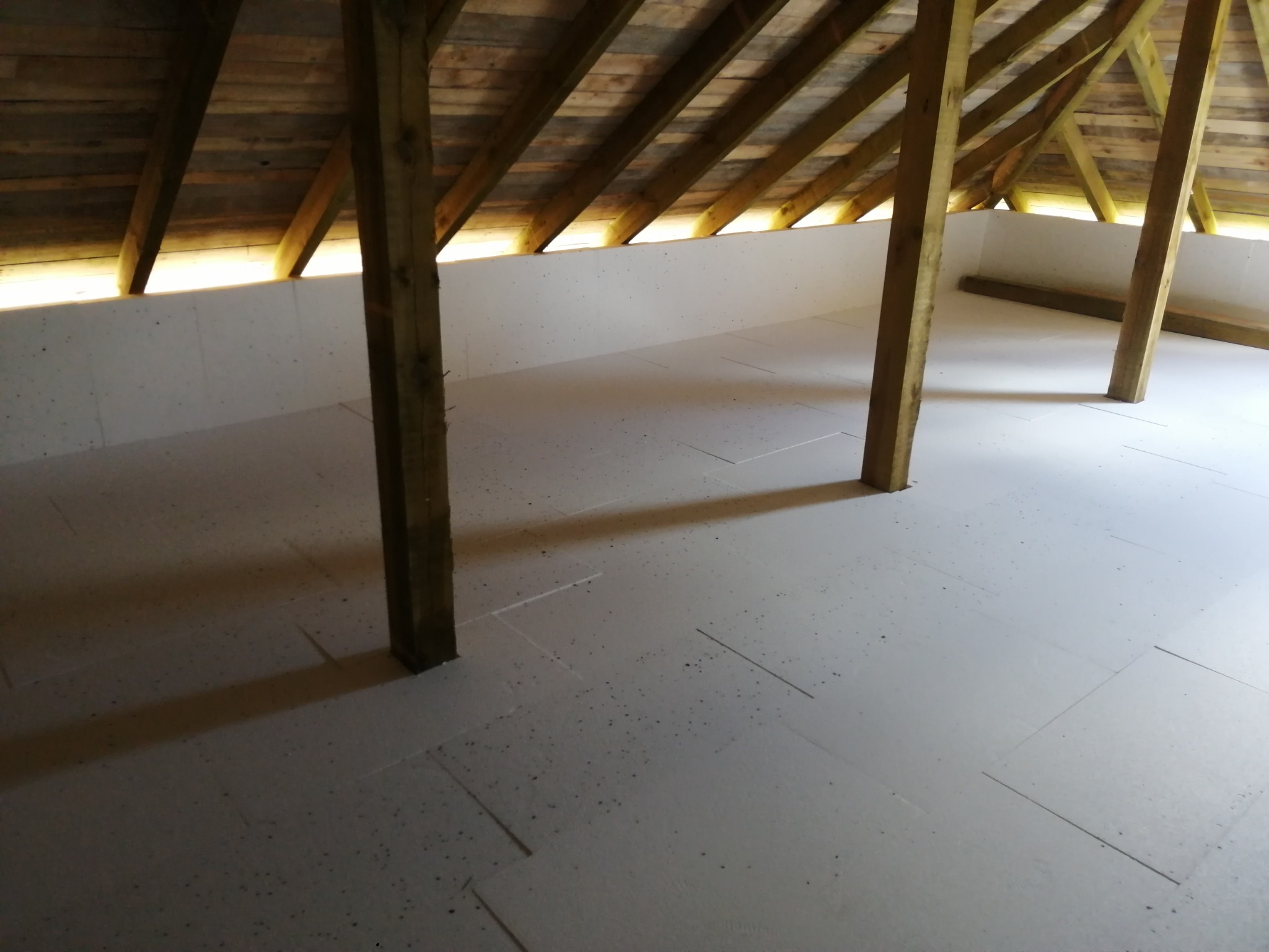 Izolacja poddasza z drewnianą konstrukcją dachu i białymi płytami izolacyjnymi na podłodze, wspartymi na drewnianych belkach, w jasnym oświetleniu.