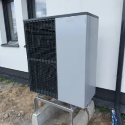 Zamontowana szara pompa ciepła Viessmann na betonowej podstawie przy białej ścianie budynku z oknami. Widoczny fragment rury spustowej.