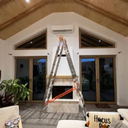 Wnętrze domu z drewnianym stropem. Na ścianie zamontowany klimatyzator, pod nim kominek. Rozstawiona drabina Krause. Widoczne okna i drzwi balkonowe.