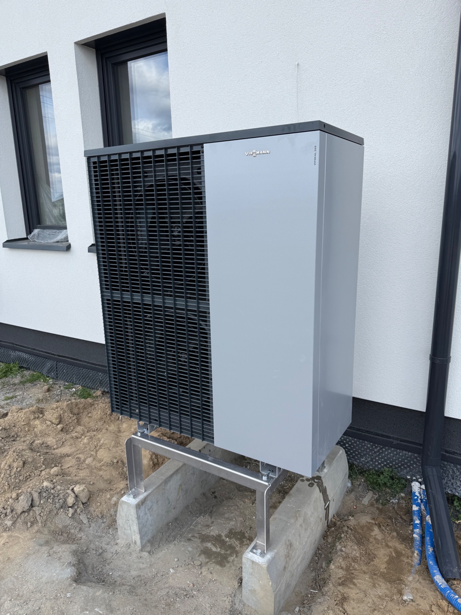 Zamontowana szara pompa ciepła Viessmann na betonowej podstawie przy białej ścianie budynku z oknami. Widoczny fragment rury spustowej.