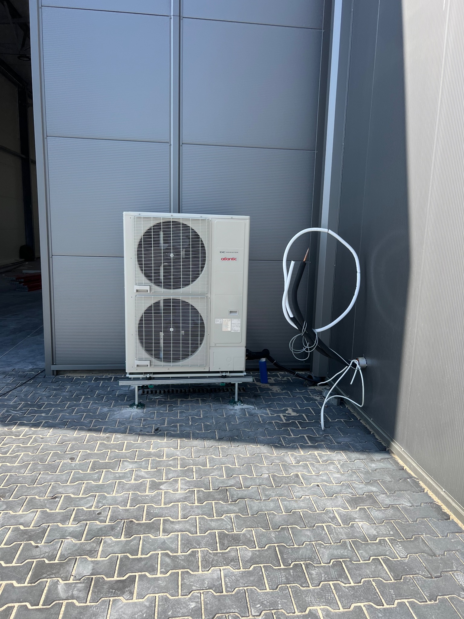 Biała, dwuczęściowa jednostka zewnętrzna klimatyzacji Atlantic na szarym tle ściany z blachy trapezowej. Widoczne rurki instalacyjne i kostka brukowa.