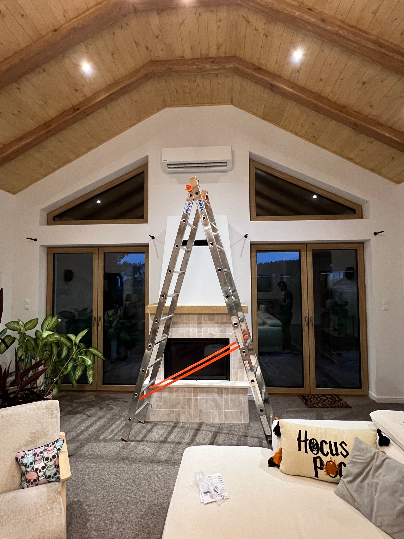 Wnętrze domu z drewnianym stropem. Na ścianie zamontowany klimatyzator, pod nim kominek. Rozstawiona drabina Krause. Widoczne okna i drzwi balkonowe.