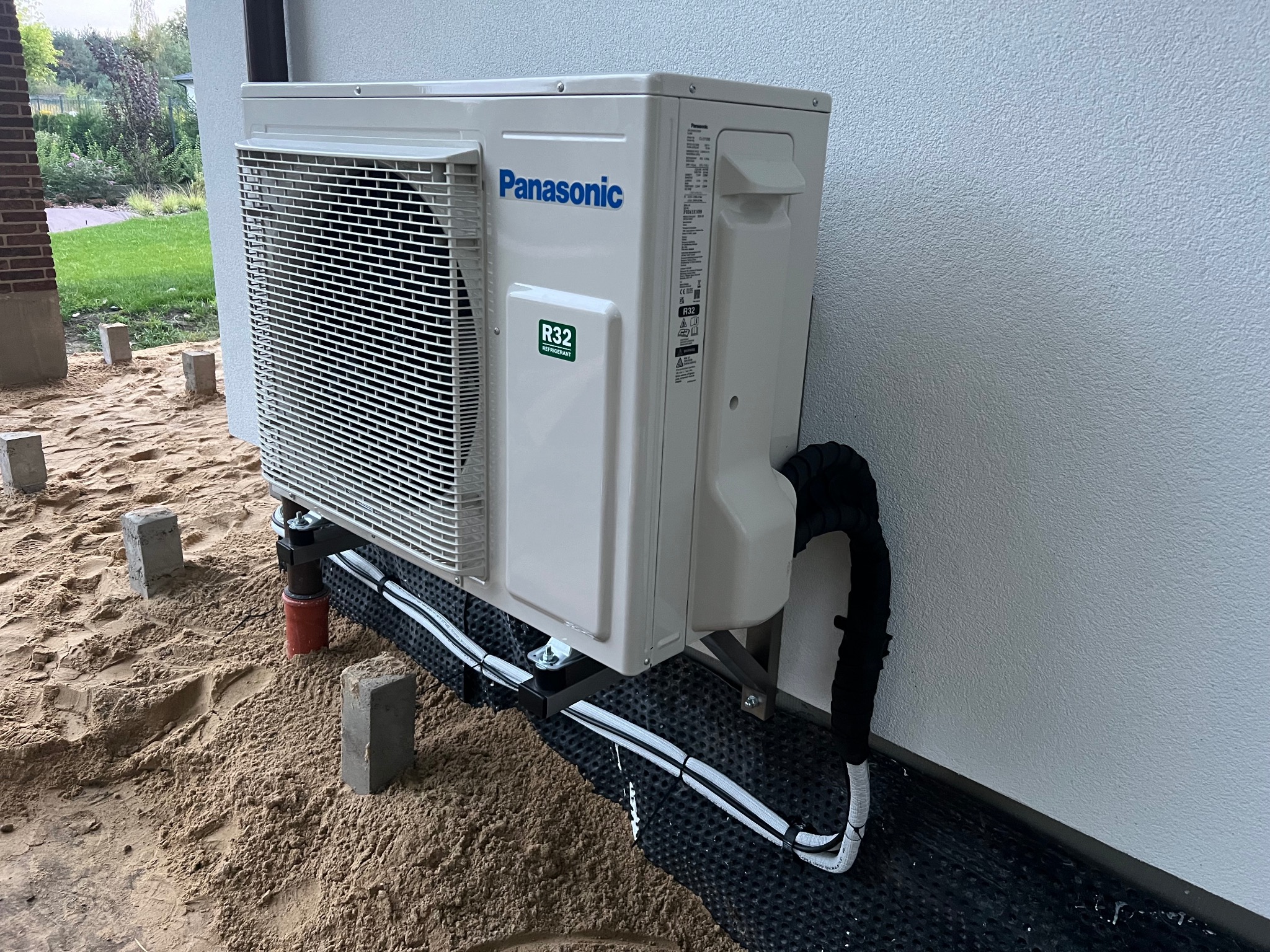 Biała, zewnętrzna jednostka klimatyzacji Panasonic R32 zamontowana na wspornikach przy ścianie budynku, z widocznymi przewodami i elementami instalacji na gruncie.