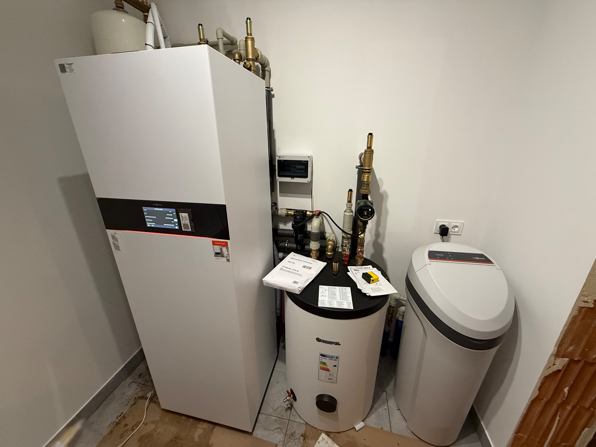 Biała pompa ciepła Viessmann, zasobnik ciepłej wody Kospel i zmiękczacz wody Viessmann w kotłowni. Widoczne rury, zawory i dokumentacja techniczna.