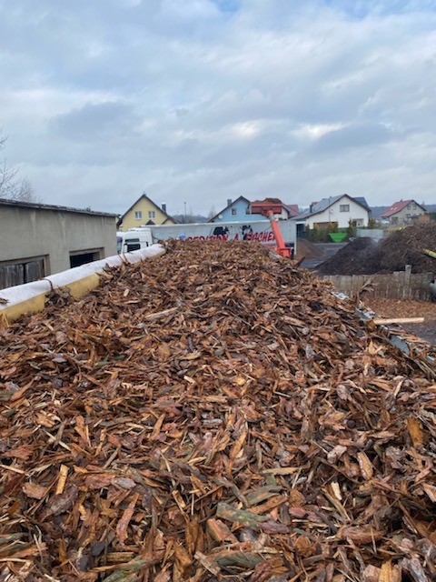 Kora drzewna w dużej pryzmie, prawdopodobnie przygotowana do transportu lub dalszego wykorzystania w ogrodnictwie, z budynkami w tle.