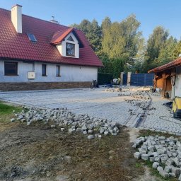 Piotr - Trwa układanie kostki granitowej na podjeździe posesji. Część powierzchni wyłożona, reszta w trakcie prac. Widoczny dom z czerwonym dachem i ogrodzenie.