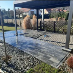 Nowoczesna pergola z utwardzoną ścieżką z kostki brukowej w odcieniach szarości, otoczona ozdobnym żwirem i roślinnością. Widoczny fragment ażurowego ogrodzenia.