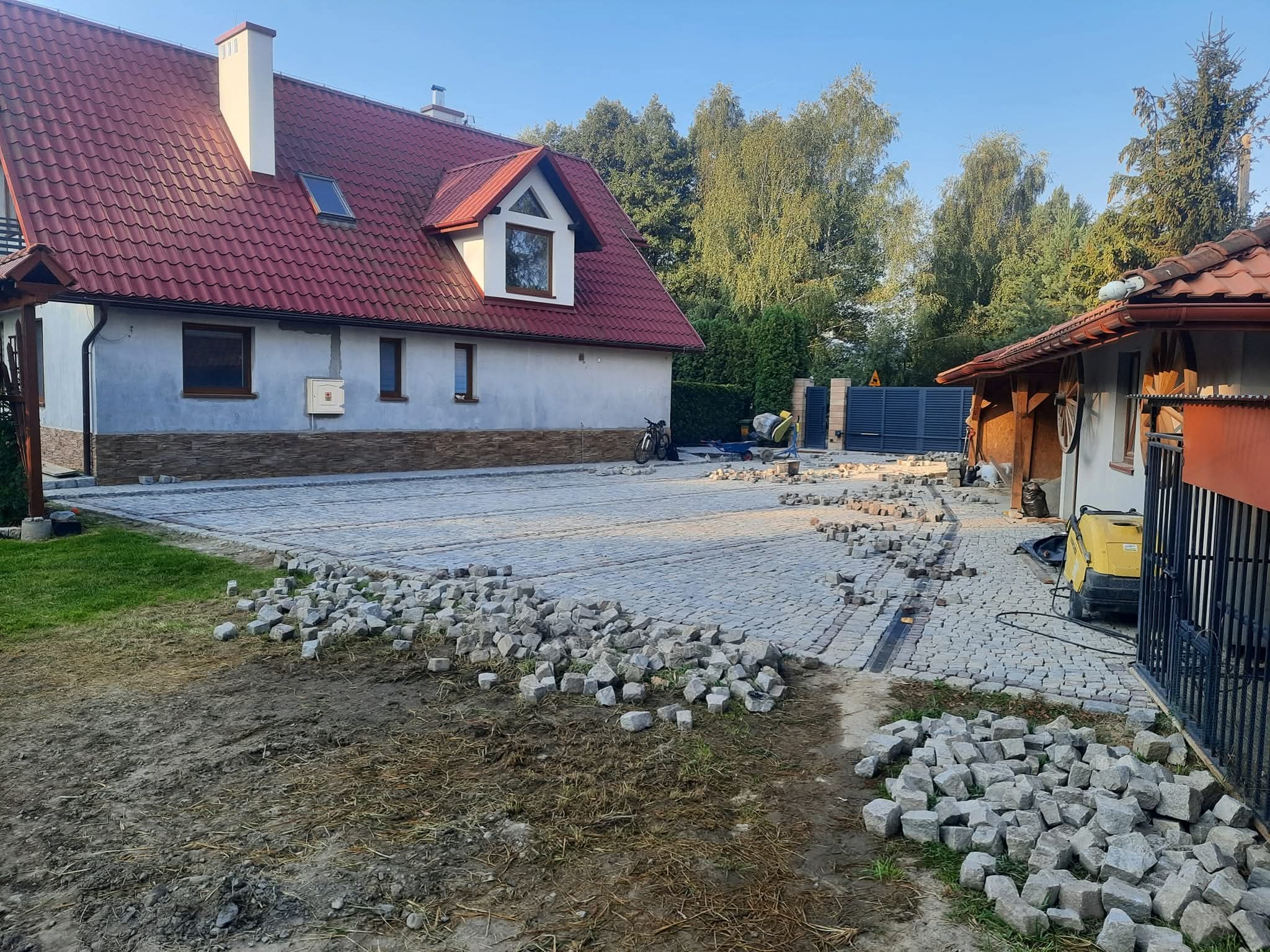 Trwa układanie kostki granitowej na podjeździe posesji. Część powierzchni wyłożona, reszta w trakcie prac. Widoczny dom z czerwonym dachem i ogrodzenie.