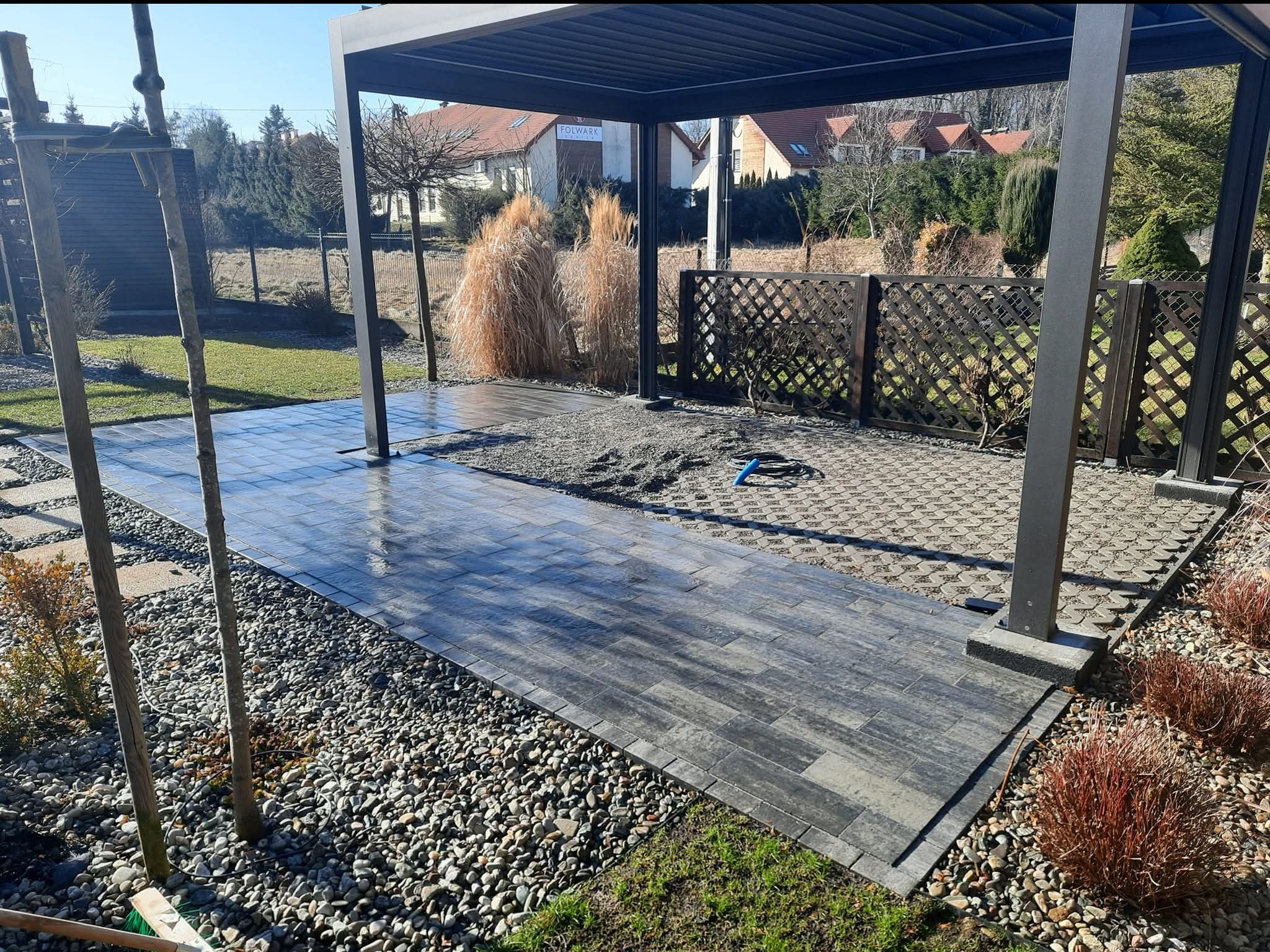 Nowoczesna pergola z utwardzoną ścieżką z kostki brukowej w odcieniach szarości, otoczona ozdobnym żwirem i roślinnością. Widoczny fragment ażurowego ogrodzenia.