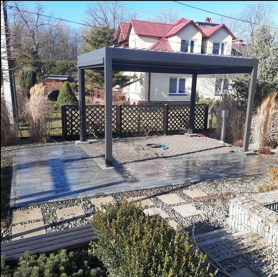 Nowoczesna pergola z szarą konstrukcją na kostce brukowej w ogrodzie, z widocznym ażurowym ogrodzeniem i domem w tle. Świeżo ułożona kostka, detale wykończenia.