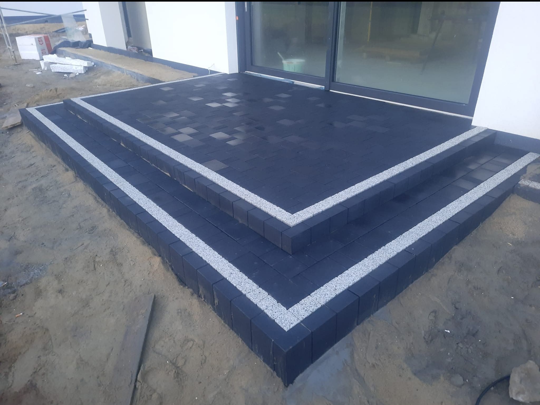 Ułożone, czarne kostki brukowe na schodach tarasu z obramowaniem z jasnego kamienia. Widoczne przesuwne drzwi tarasowe i przygotowany teren.