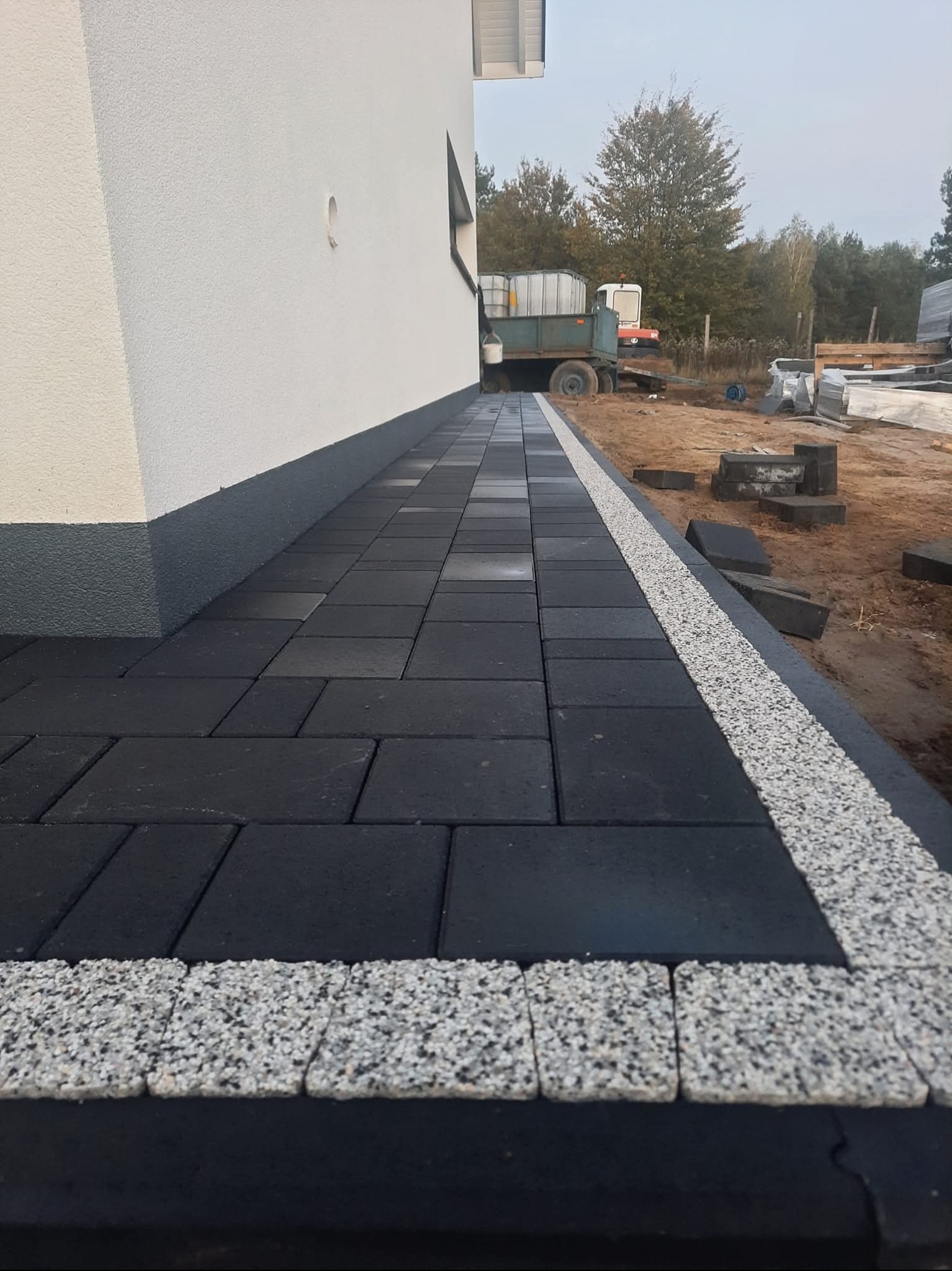 Nowoczesna ścieżka z czarnej kostki brukowej z obrzeżem z jasnego kruszywa, przylegająca do ściany budynku. W tle widoczny sprzęt budowlany.