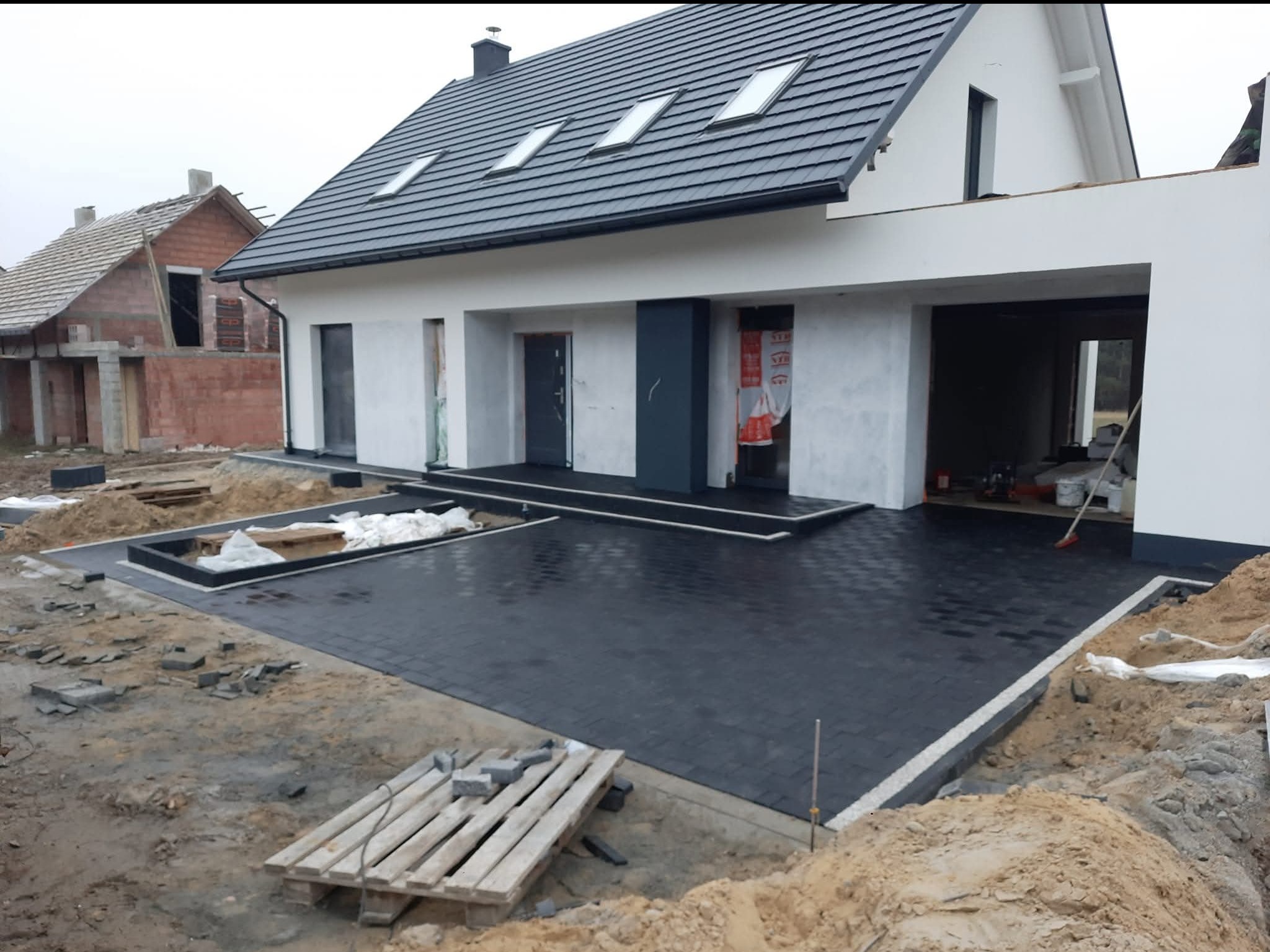 Nowoczesny dom z antracytową kostką brukową ułożoną na podjeździe. Widoczne palety z kostką i przygotowane obrzeża. Częściowo zakończone prace brukarskie.