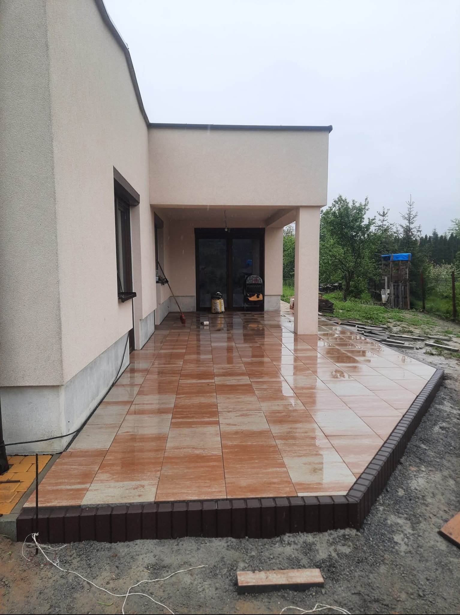 Wykończony taras z brązowej kostki brukowej przy domu, z ciemnobrązowym obrzeżem. Widoczne detale wykończenia i otoczenie w trakcie prac.