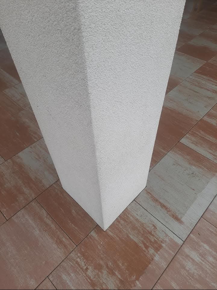 Fragment kolumny z dekoracyjnym tynkiem, stojącej na brązowo-szarej kostce brukowej. Widoczny narożnik kolumny i wzór kostki.