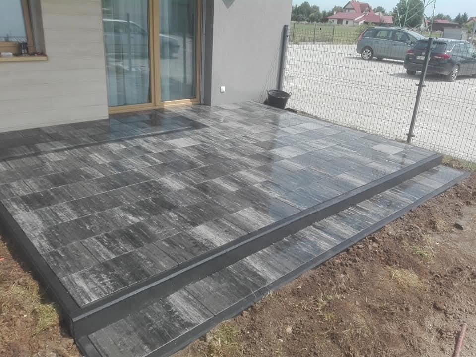 Ułożona kostka brukowa w odcieniach szarości tworząca taras z dwoma stopniami, widok na dom z przesuwnymi drzwiami i metalowe ogrodzenie w tle.