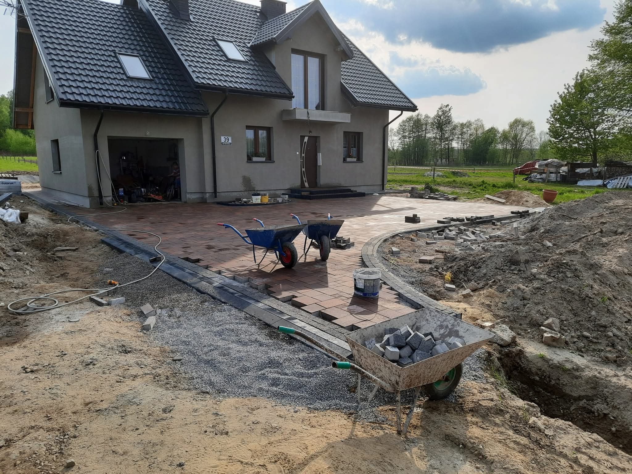 Ułożona część podjazdu z kostki brukowej przy domu z szarą elewacją i ciemnym dachem. W tle widać dwie taczki i narzędzia. Na pierwszym planie taczka z kostką granitową.