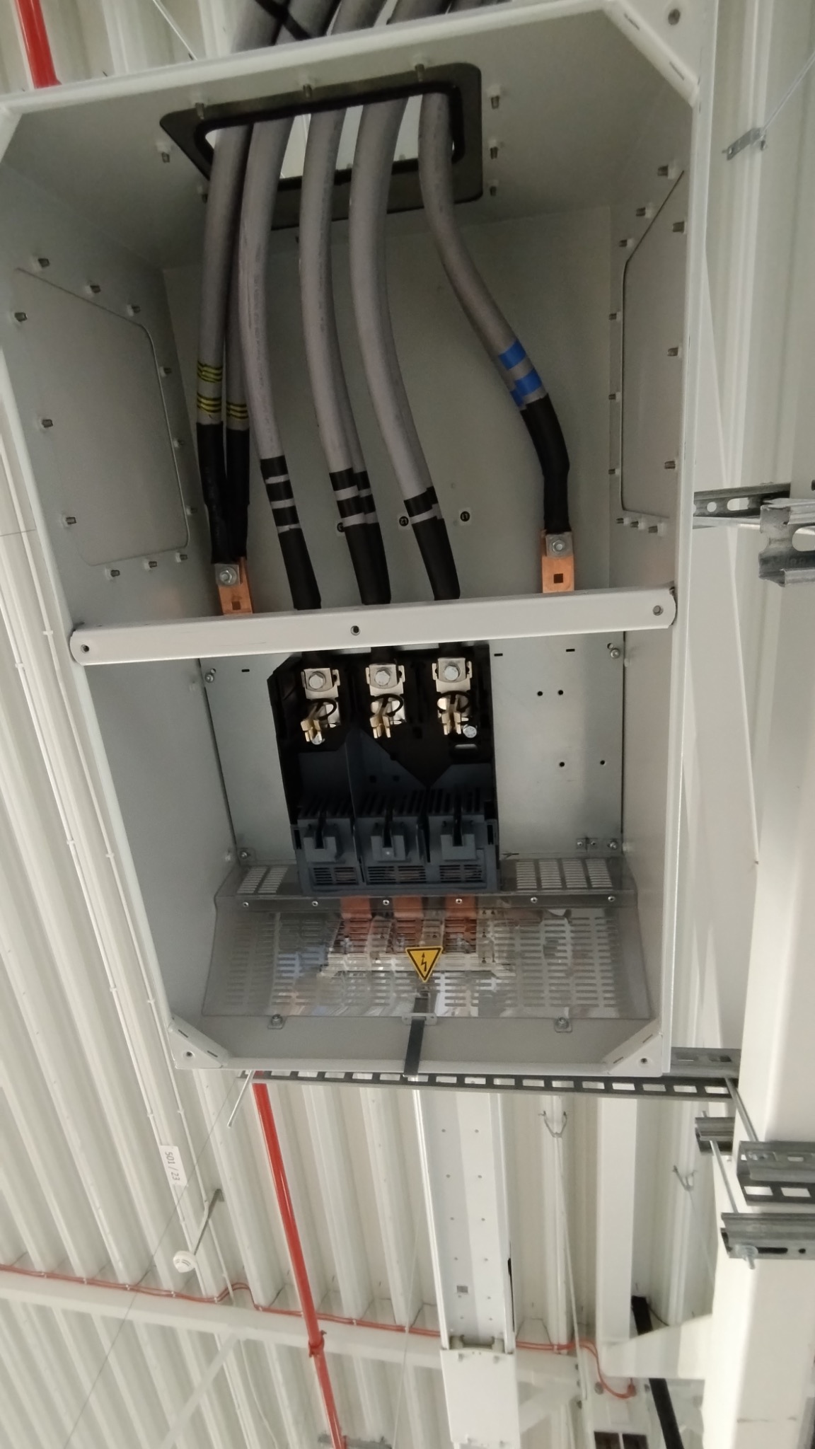 Szara skrzynka elektryczna z przewodami i złączami, zamontowana pod sufitem z blachy trapezowej. Widoczne oznaczenia na przewodach i ostrzeżenie o napięciu.