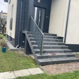 Schody wejściowe z szarą balustradą na tle nowoczesnego domu z ciemnej cegły i jasnego tynku. Na schodach leżą narzędzia.