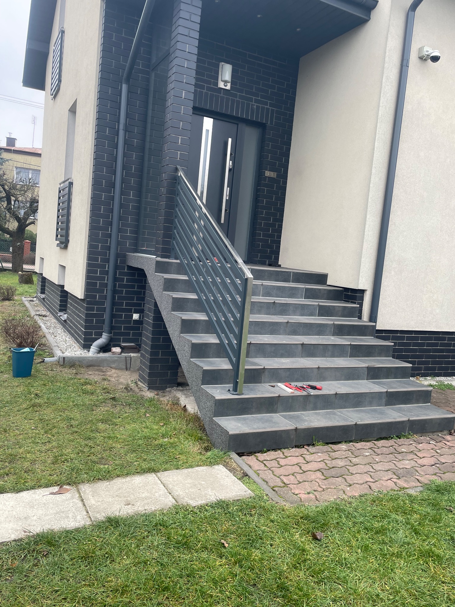 Schody wejściowe z szarą balustradą na tle nowoczesnego domu z ciemnej cegły i jasnego tynku. Na schodach leżą narzędzia.