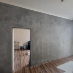 Ściana w odcieniach szarości z efektem dekoracyjnym, widok na wnętrze przez otwarte drzwi, drewniana podłoga, minimalistyczny design.