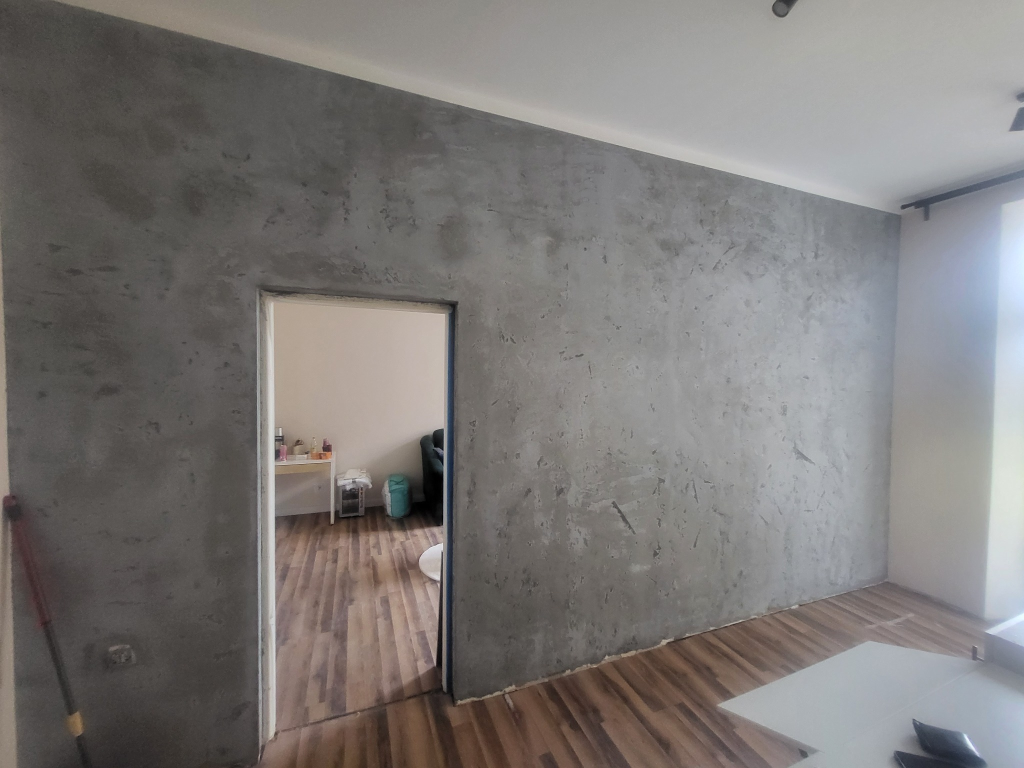 Ściana w odcieniach szarości z efektem dekoracyjnym, widok na wnętrze przez otwarte drzwi, drewniana podłoga, minimalistyczny design.