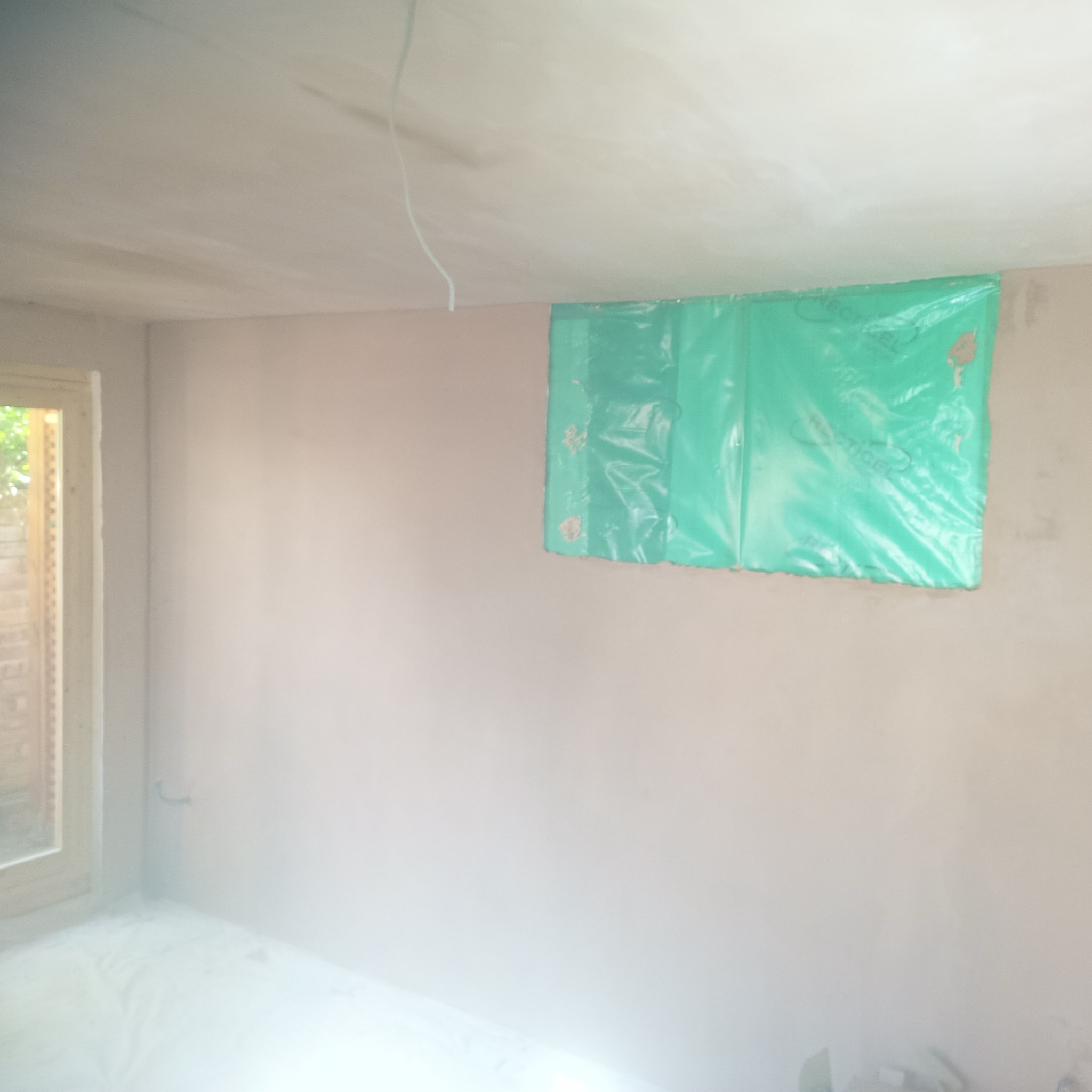 Wnętrze po szpachlowaniu ścian i sufitu, zabezpieczone okno folią. Widoczne gładzie na ścianach i przygotowanie do dalszych prac wykończeniowych.