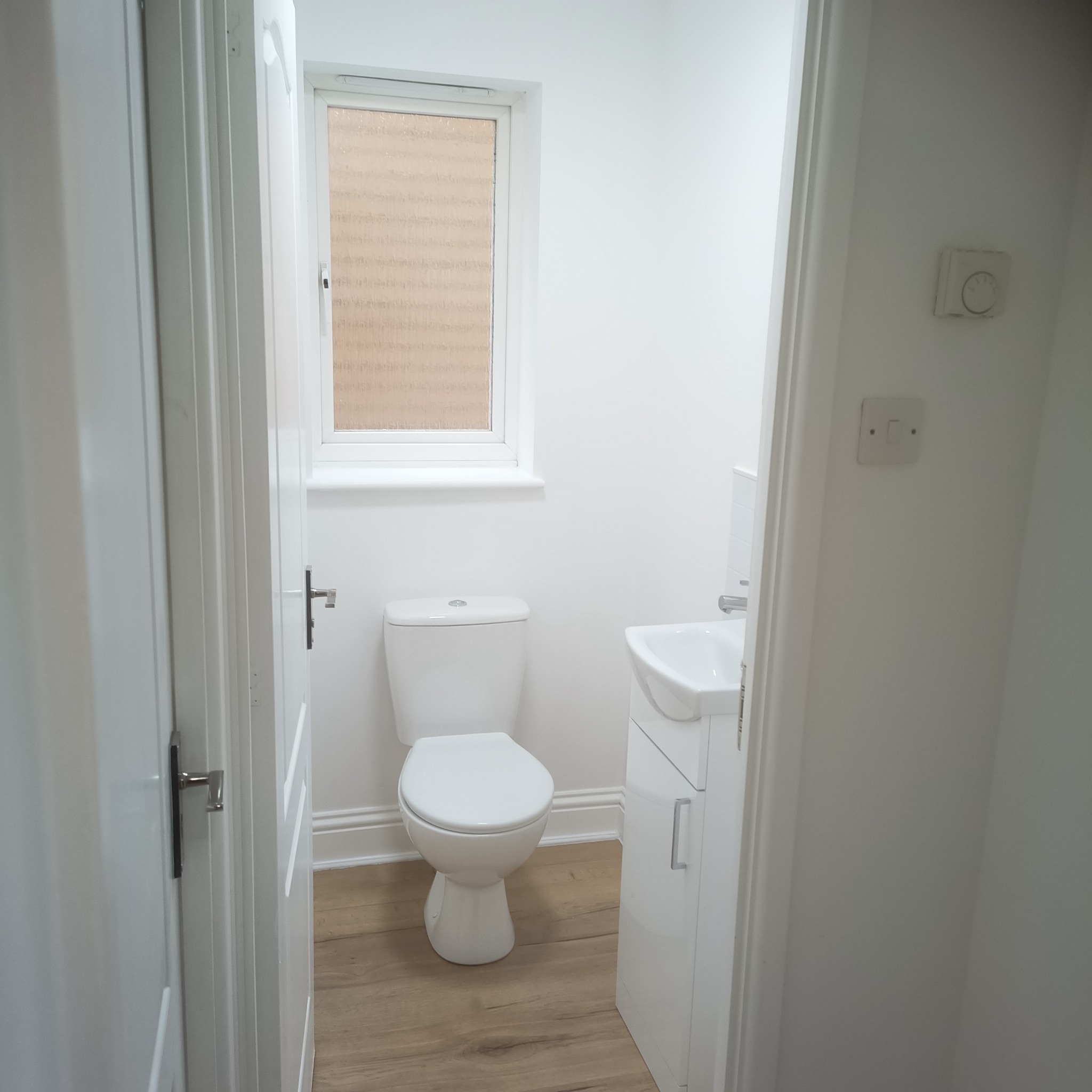 Jasna, mała łazienka po remoncie: białe ściany, toaleta, umywalka z szafką i drewnopodobna podłoga. Widok zza uchylonych drzwi.