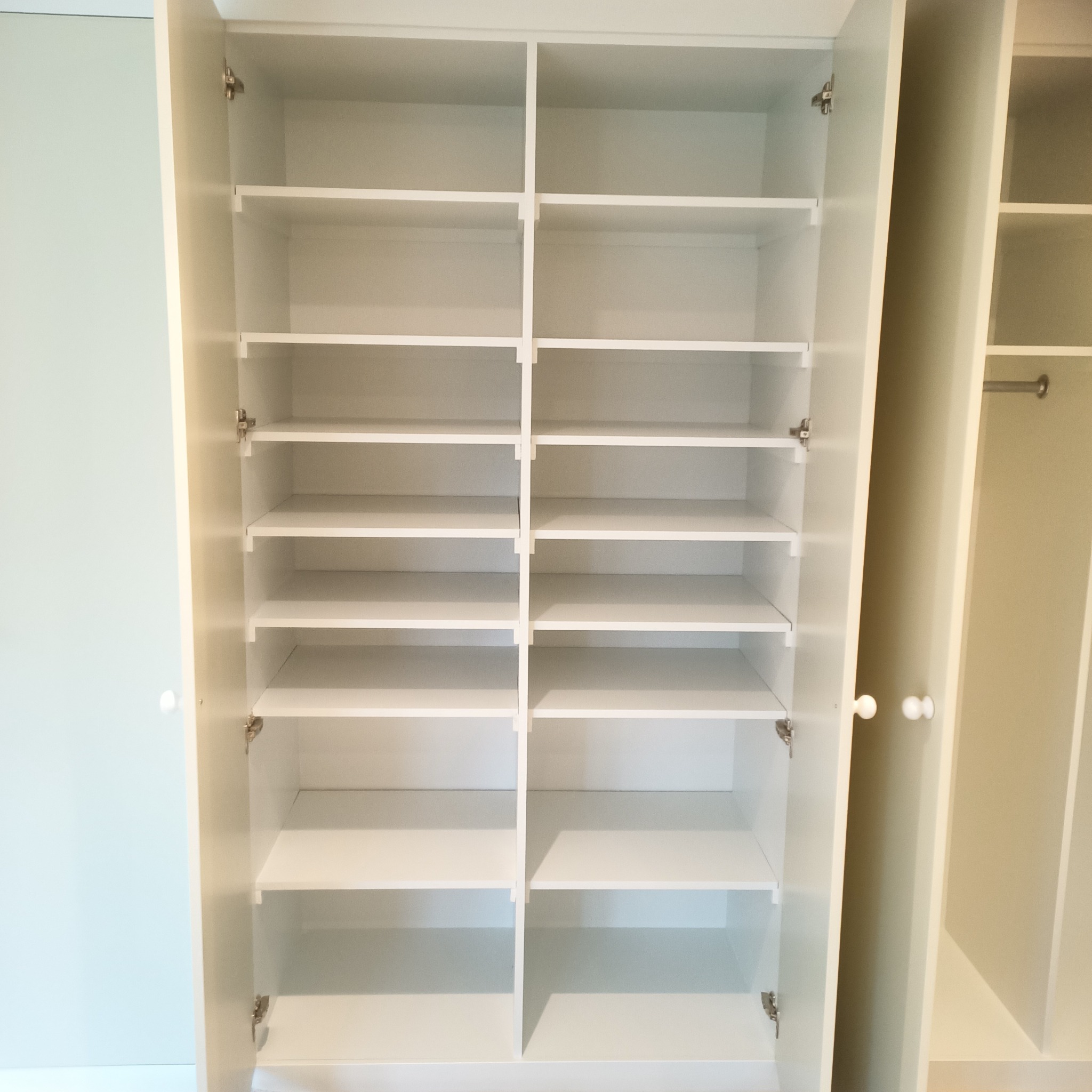 Biała, otwarta szafa z półkami, widok z przodu. Drzwi szafy po lewej stronie, na zawiasach. Półki równo rozmieszczone, puste. Elementy garderoby po prawej.