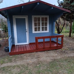 Tata synami - Niebieski domek narzędziowy z czerwonym dachem i werandą, widoczny z poziomu gruntu, otoczony zielenią ogrodu. Świeżo pomalowany.