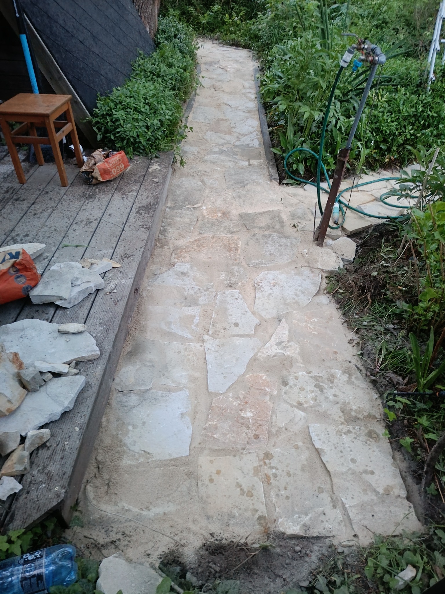 Kamienna ścieżka ogrodowa w trakcie budowy, widoczne materiały budowlane i częściowo ułożone kamienie, otoczenie zieleni i drewniany podest.