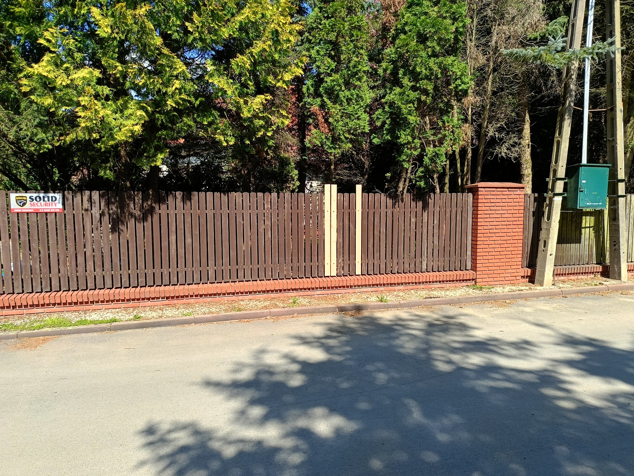 Brunatny płot z ceglaną podmurówką i słupkiem, zabezpieczony przez Solid Security. Fragment płotu w surowym drewnie. Zadrzewione tło, cień na asfalcie.